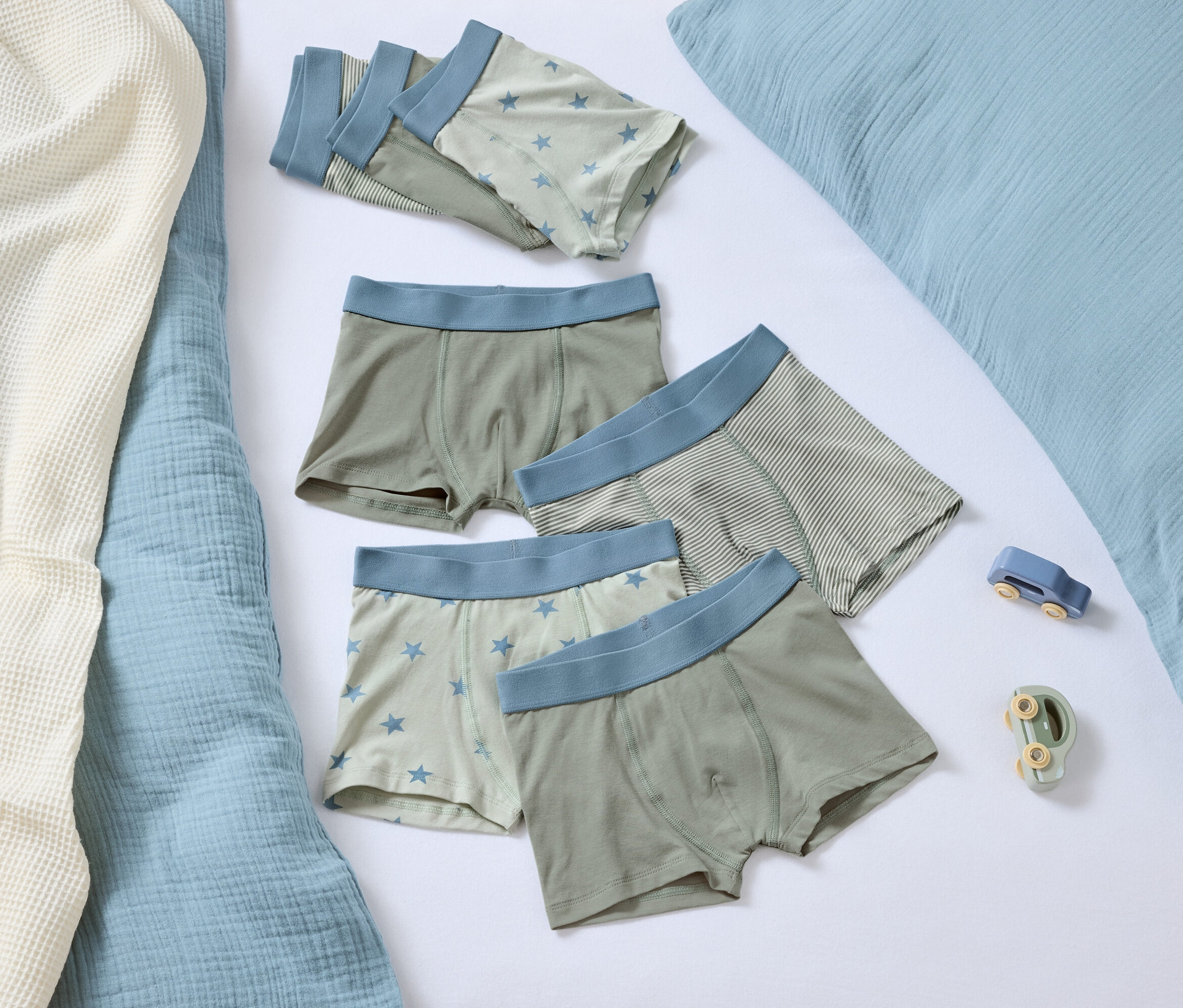 Cinq boxers avec une bordure bleue reposent sur un fond blanc à côté d'un oreiller bleu et de deux petits jouets.