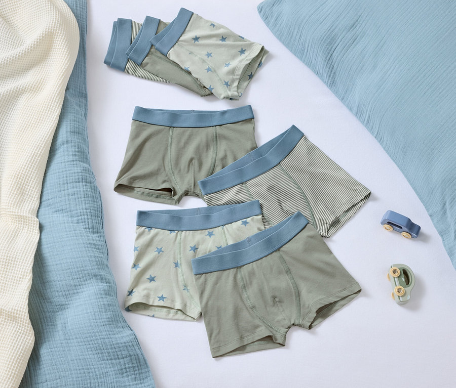 Cinq boxers avec une bordure bleue reposent sur un fond blanc à côté d'un oreiller bleu et de deux petits jouets.
