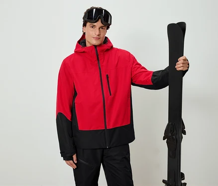 Un homme portant une veste rouge et noire tient des skis noirs et porte des lunettes de ski.