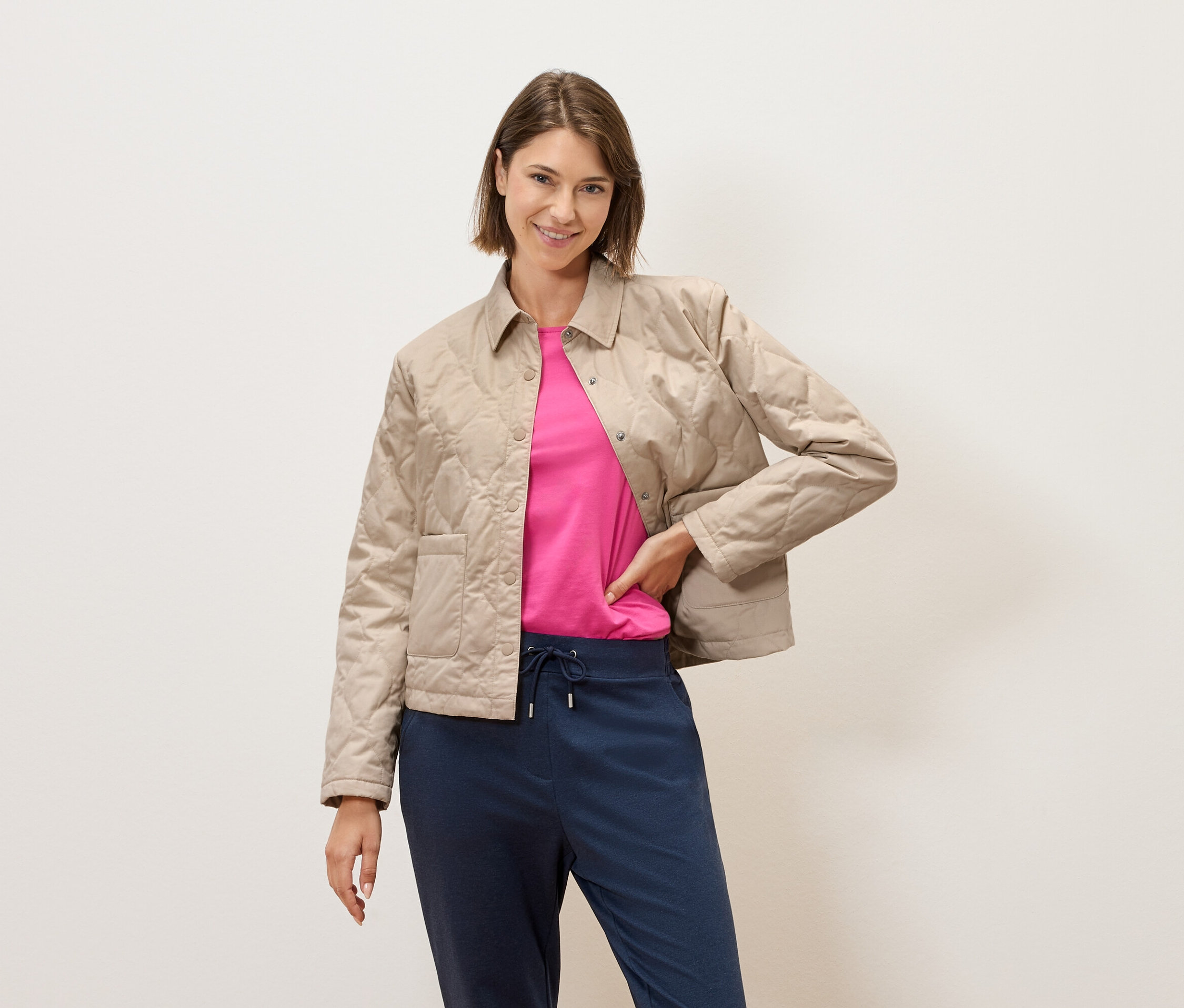 Une femme aux cheveux bruns courts pose devant un mur crème, portant une veste matelassée beige, un t-shirt rose et un pantalon bleu.