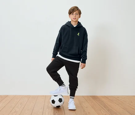 Un garçon se tient un pied sur un ballon de football, portant un sweat à capuche bleu marine et un pantalon noir.