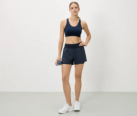 Une femme se tient debout, la main sur la hanche, portant un soutien-gorge de sport bleu marine, un short et des baskets blanches, tenant une bouteille d'eau.