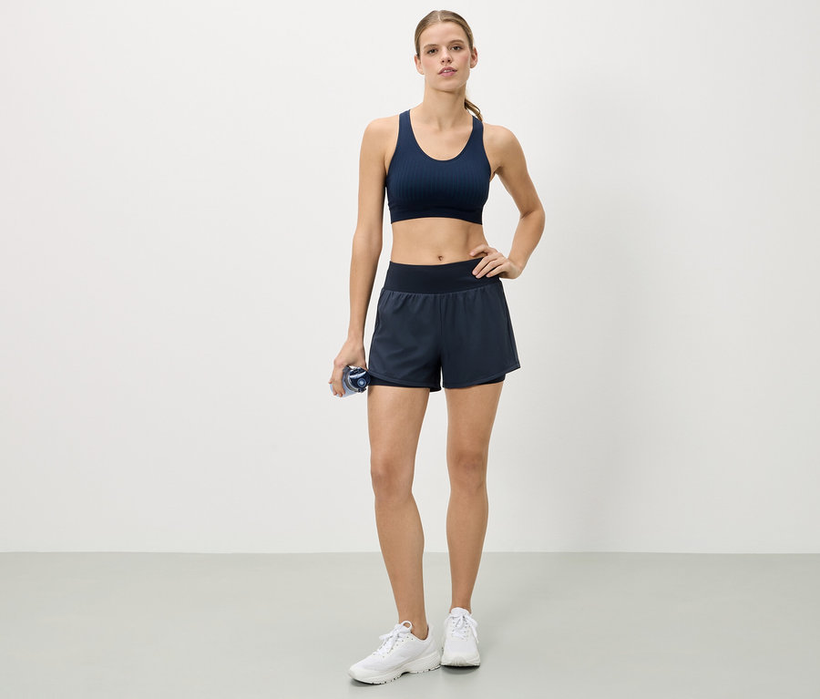 Une femme se tient debout, la main sur la hanche, portant un soutien-gorge de sport bleu marine, un short et des baskets blanches, tenant une bouteille d'eau.