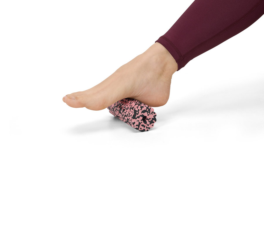 Un pied en legging bordeaux roule sur un rouleau de massage rose et noir moucheté sur fond blanc.