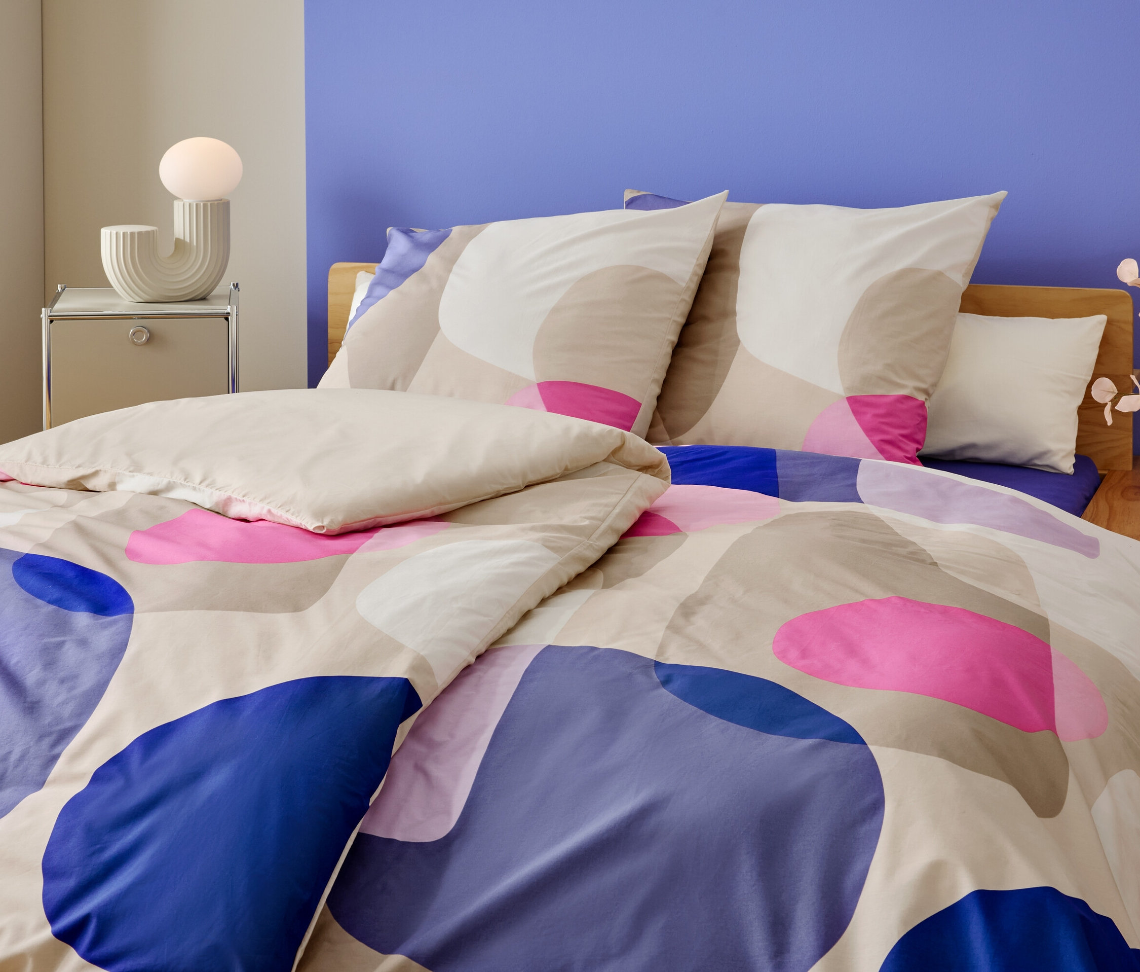 Gros plan d'un lit avec un motif géométrique bleu, rose et beige. Une lampe et un vase de fleurs sont posés sur la table de chevet.