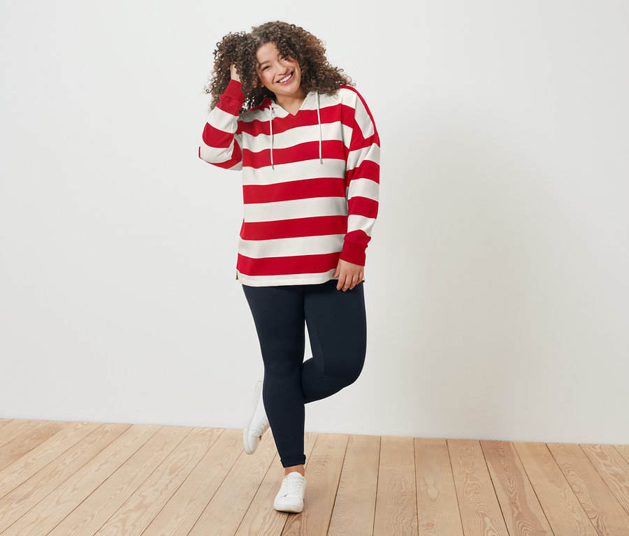 Femme aux cheveux bouclés portant un haut rayé rouge et blanc et un legging bleu debout sur un plancher en bois.