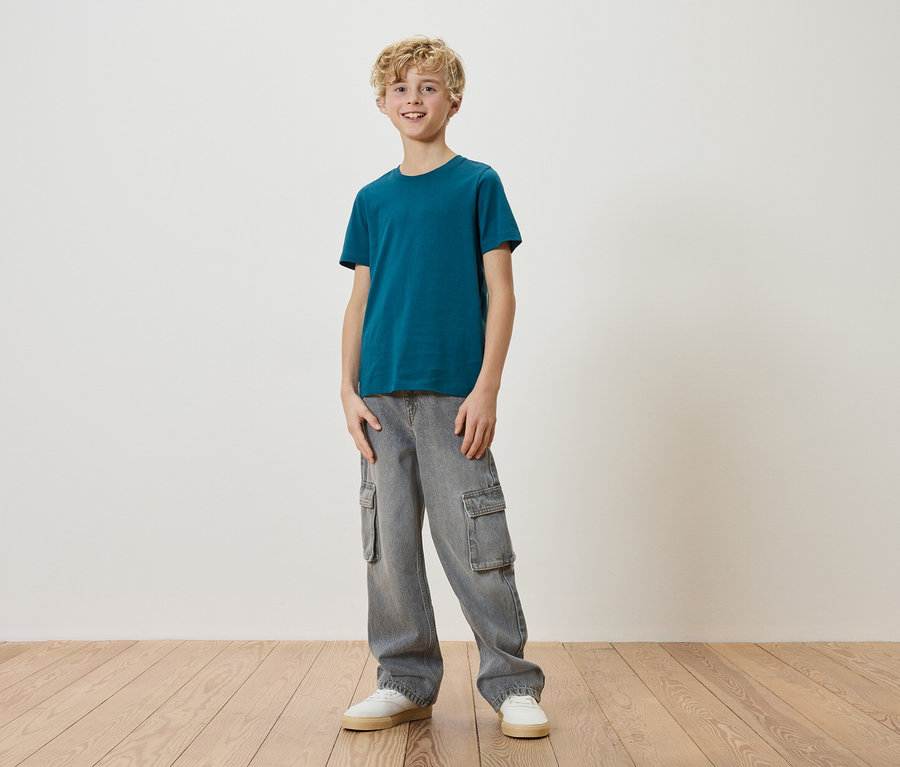 Un garçon blond se tient devant un mur blanc, vêtu d'un t-shirt turquoise, d'un pantalon cargo gris et de baskets blanches.