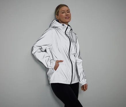 Une femme, la main dans la poche, porte une veste réfléchissante à capuche et un legging noir sur un fond gris.
