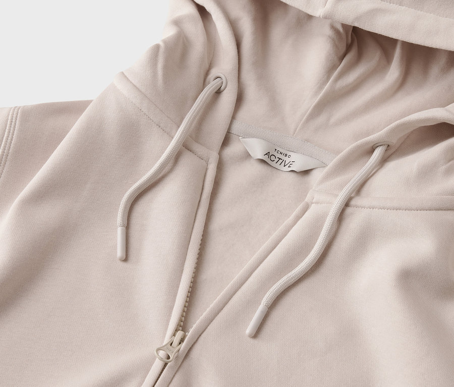 Gros plan d'un sweat à capuche beige clair avec fermeture éclair et cordon de serrage. Une étiquette indiquant « TCHIBO ACTIVE » est visible.