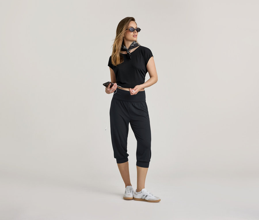 Une femme portant un haut noir et un pantalon 3/4 de yoga tient un téléphone et des écouteurs. Elle porte un foulard et des baskets.