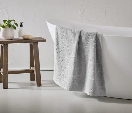 Une serviette grise à motifs géométriques est suspendue au bord d'une baignoire blanche. Un tabouret en bois se trouve à côté, sur lequel sont posés une plante, du savon et une éponge.