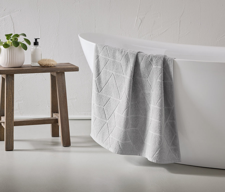 Une serviette grise à motifs géométriques est suspendue au bord d'une baignoire blanche. Un tabouret en bois se trouve à côté, sur lequel sont posés une plante, du savon et une éponge.
