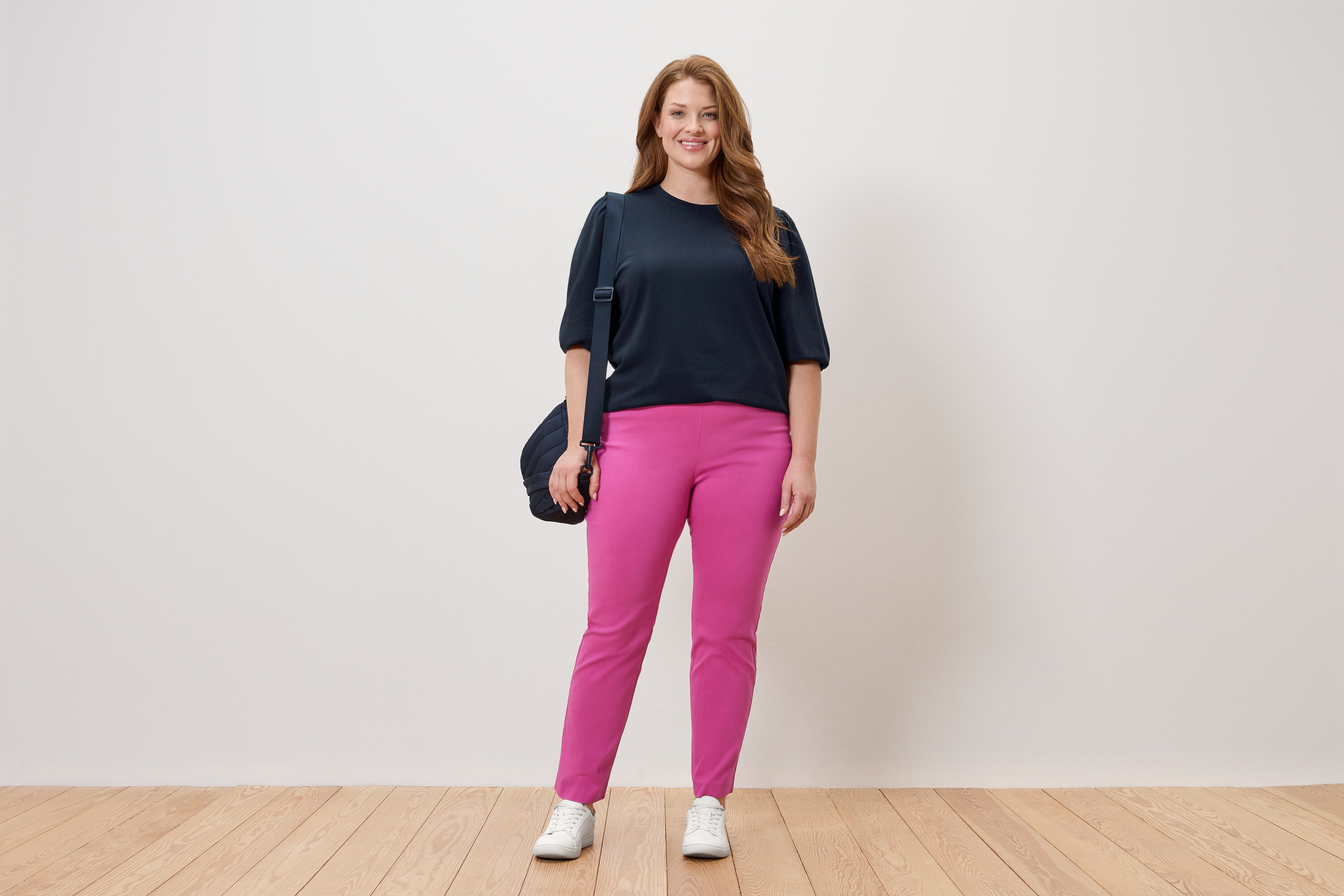 Femme debout en haut bleu marine et pantalon rose avec un sac à bandoulière devant un mur blanc.