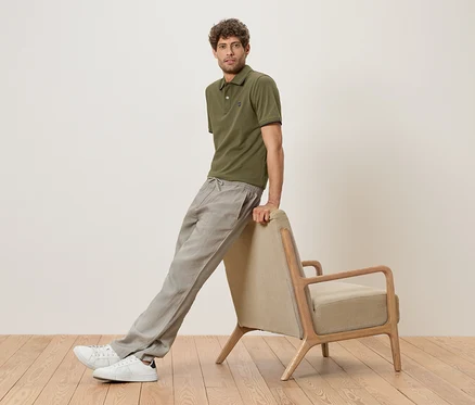 Un homme est appuyé contre un fauteuil beige, portant un polo vert olive, un pantalon en lin gris et des baskets blanches.