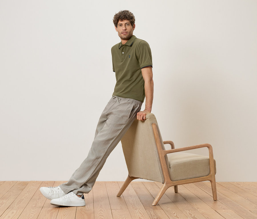 Un homme est appuyé contre un fauteuil beige, portant un polo vert olive, un pantalon en lin gris et des baskets blanches.
