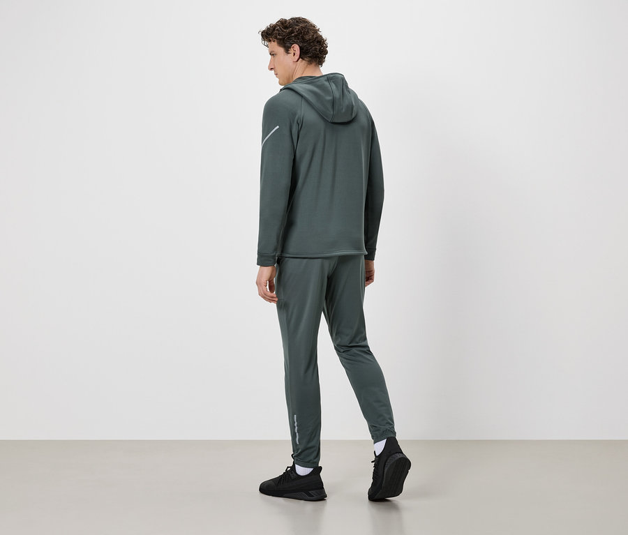 Homme de dos portant un survêtement vert foncé à capuche, des chaussures noires et des chaussettes blanches sur fond blanc.
