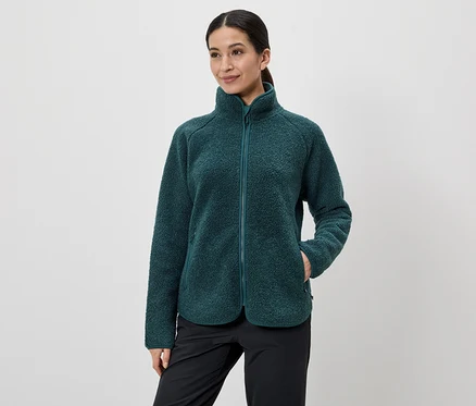 Une femme aux cheveux attachés en queue de cheval porte une veste en polaire verte zippée et un pantalon noir, une main dans la poche de sa veste.