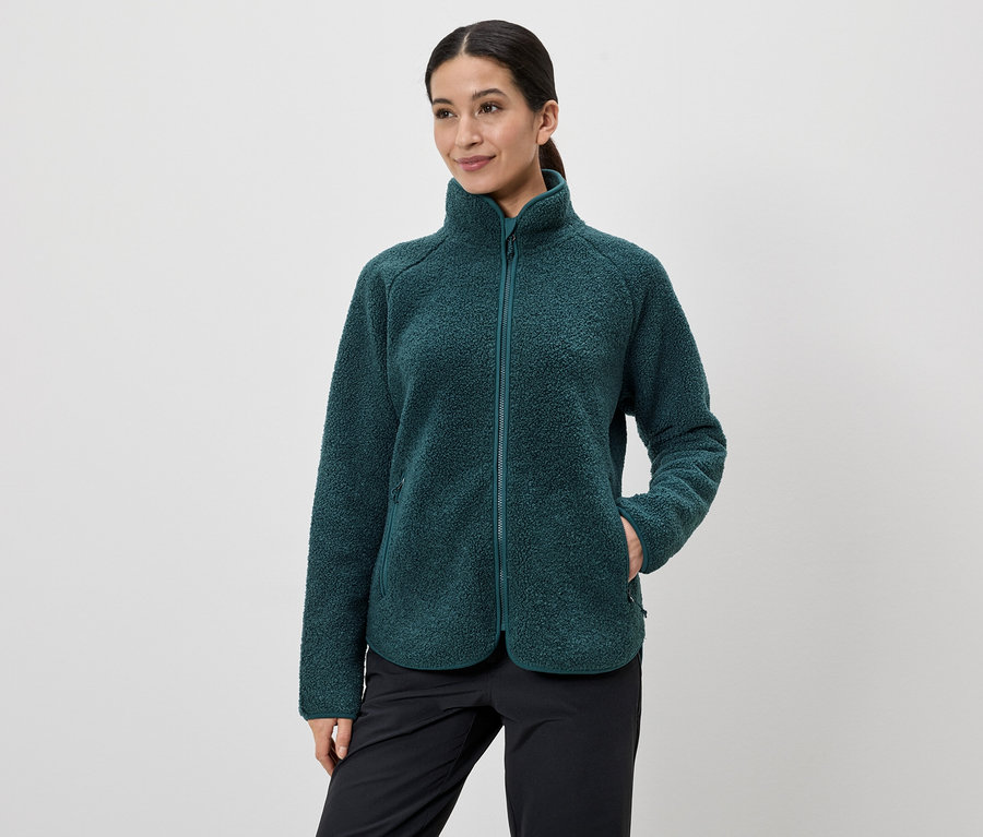 Une femme aux cheveux attachés en queue de cheval porte une veste en polaire verte zippée et un pantalon noir, une main dans la poche de sa veste.
