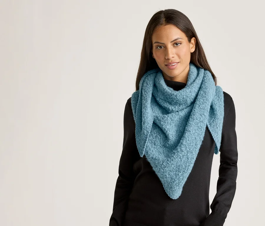 Une femme aux longs cheveux foncés porte un châle triangulaire bleu clair et un pull noir sur un fond clair.