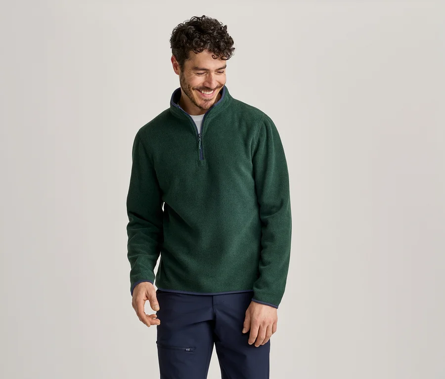 Un homme souriant aux cheveux foncés et à la barbe porte un haut en polaire vert foncé avec un quart de zip et un pantalon bleu foncé.