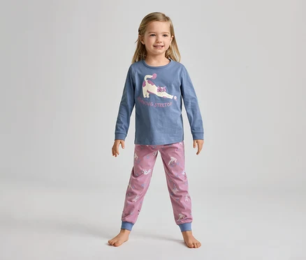 Une jeune fille souriante, pieds nus, porte un haut bleu à manches longues avec un chat qui s'étire et un pantalon rose à motifs de licornes.