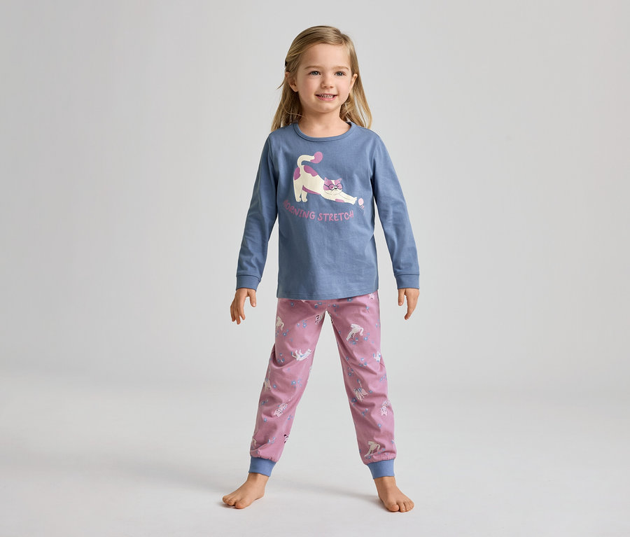 Une jeune fille souriante, pieds nus, porte un haut bleu à manches longues avec un chat qui s'étire et un pantalon rose à motifs de licornes.