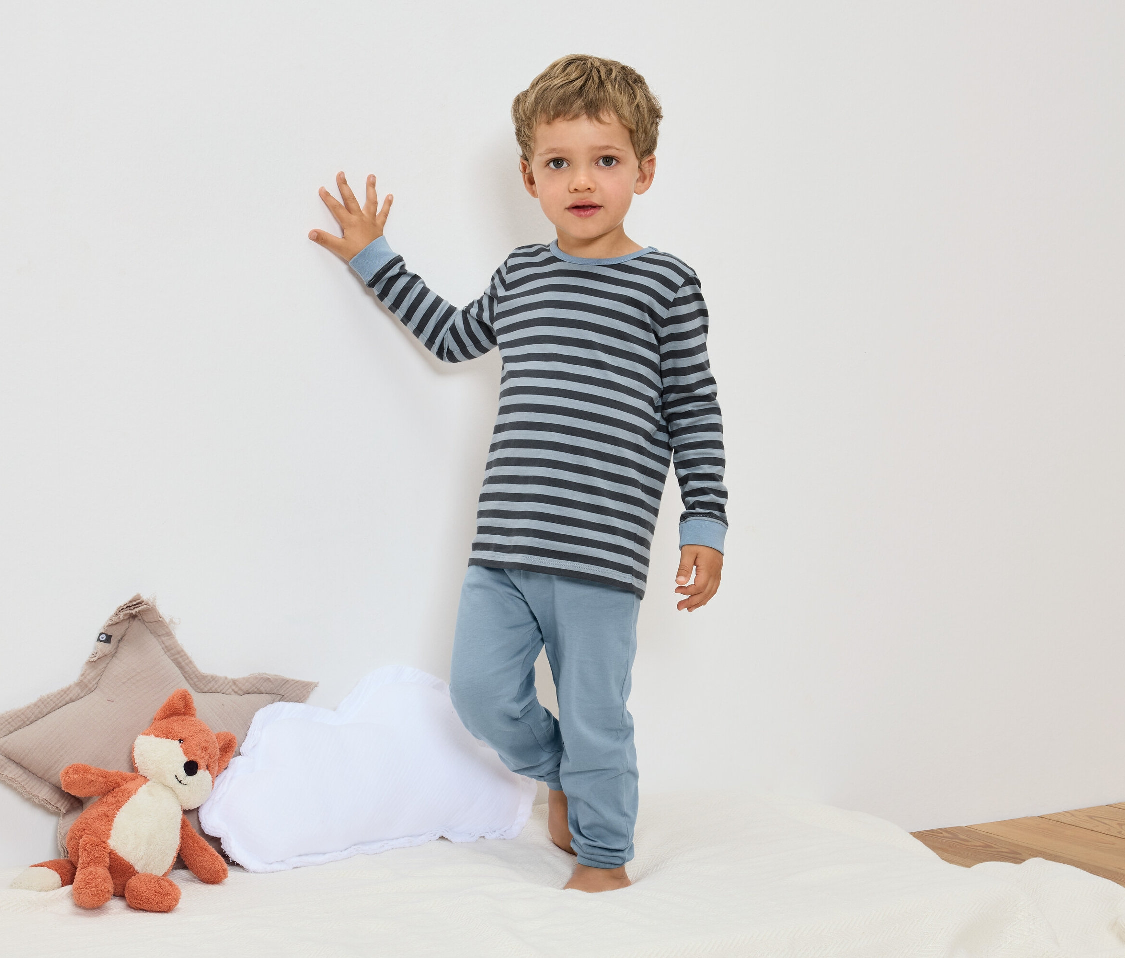 Un garçon se tient en pyjama rayé bleu à côté d'une peluche renard et d'oreillers.