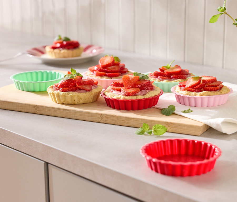 Plusieurs tartelettes aux fraises dans des moules colorés disposées sur une planche à découper en bois, garnies de menthe.