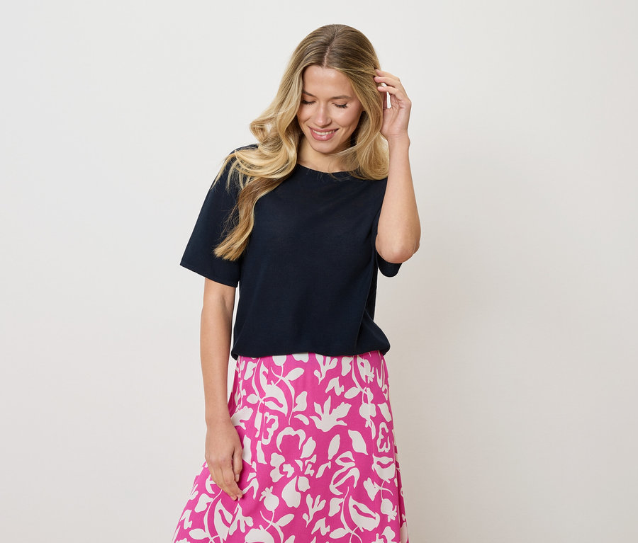 Femme aux cheveux blonds ondulés portant un t-shirt bleu marine et une jupe rose et blanche à motif floral, touchant ses cheveux d'une main.