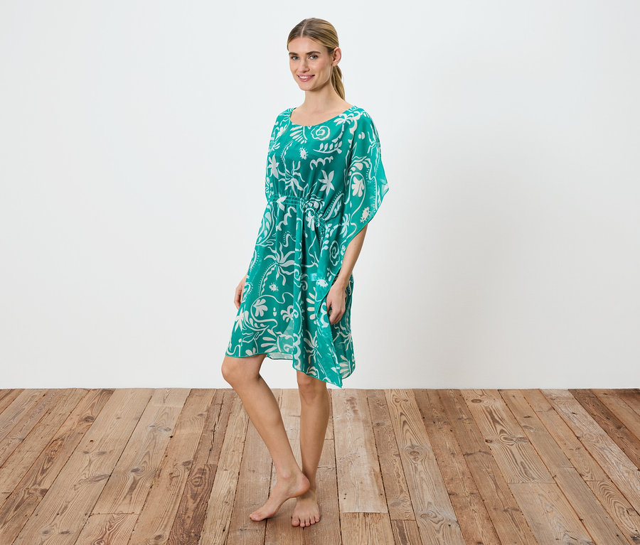 Une femme se tient pieds nus sur un plancher en bois, portant une robe verte avec un motif floral blanc et un cordon de serrage à la taille.