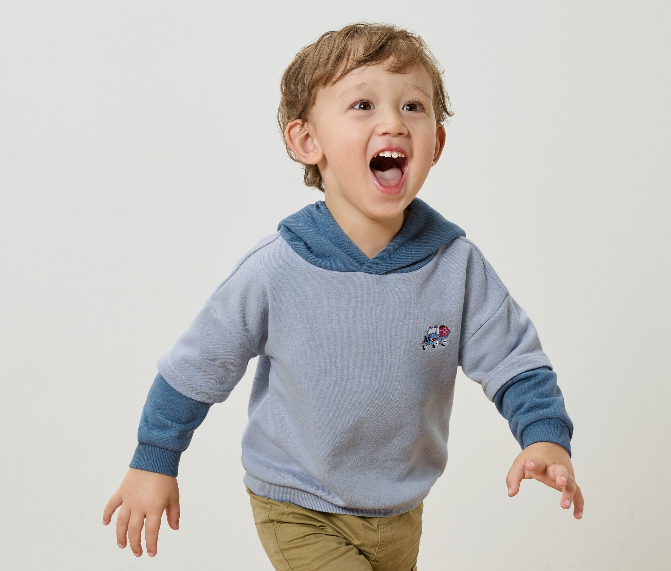 Un petit garçon aux boucles blondes et à la bouche ouverte porte un sweat à capuche bleu clair avec un petit appliqué de voiture et un pantalon marron clair sur un fond blanc.