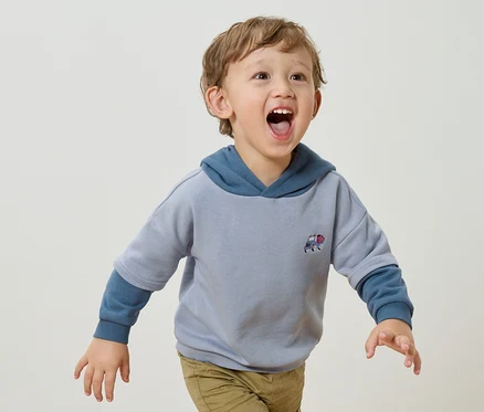 Un petit garçon aux boucles blondes et à la bouche ouverte porte un sweat à capuche bleu clair avec un petit appliqué de voiture et un pantalon marron clair sur un fond blanc.