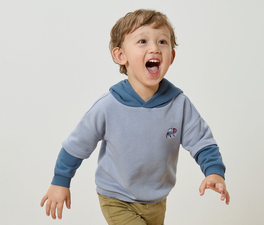 Un petit garçon aux boucles blondes et à la bouche ouverte porte un sweat à capuche bleu clair avec un petit appliqué de voiture et un pantalon marron clair sur un fond blanc.