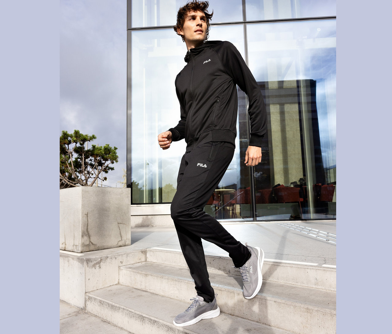 Homme en survêtement Fila noir descendant un escalier en béton, chaussures Fila grises.