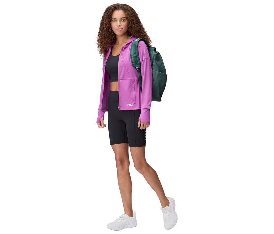 Une femme aux cheveux bouclés porte une veste Fila violette, un short noir, un crop top noir, des baskets blanches et un sac à dos vert.
