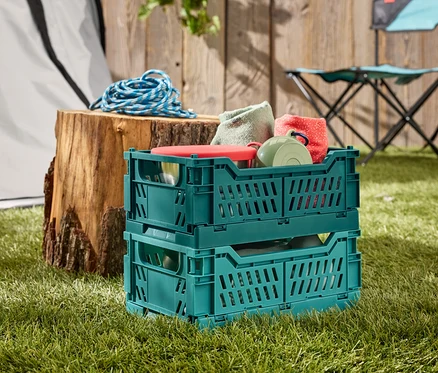 Deux caisses pliantes vertes empilées sur l'herbe, remplies de vaisselle et de textiles. En arrière-plan, une souche d'arbre avec une corde bleue et une chaise de camping.