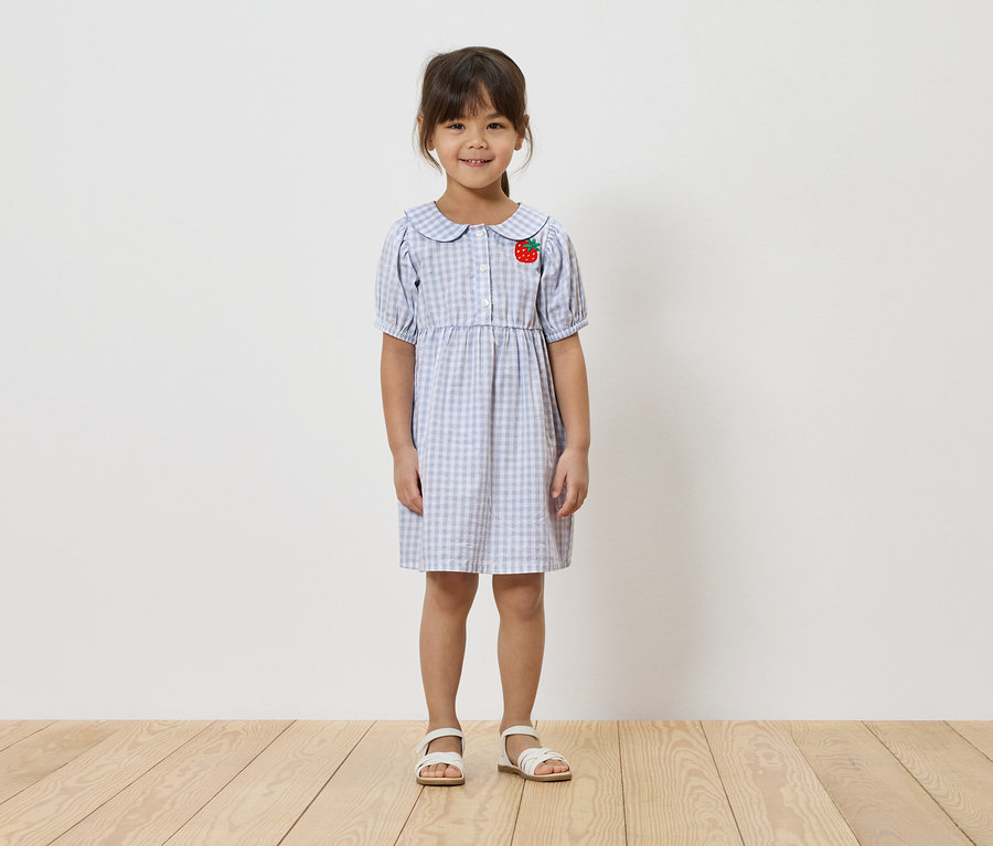 Fillette portant une robe à carreaux bleu clair avec une application fraise et des sandales blanches.