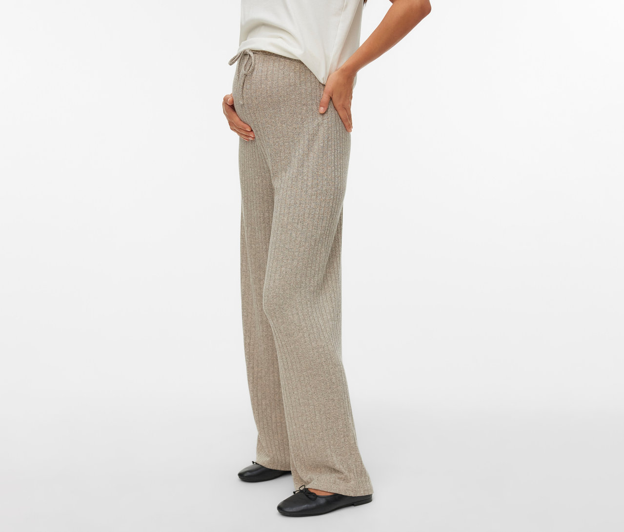 Femme enceinte portant un pantalon Loosefit MAMALICIOUS « MLPAMMIE », un t-shirt blanc et des chaussures noires, la main sur le ventre.