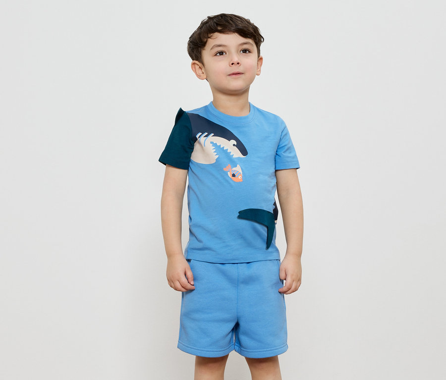 Un garçon porte un t-shirt bleu clair avec un motif de requin et un short assorti.