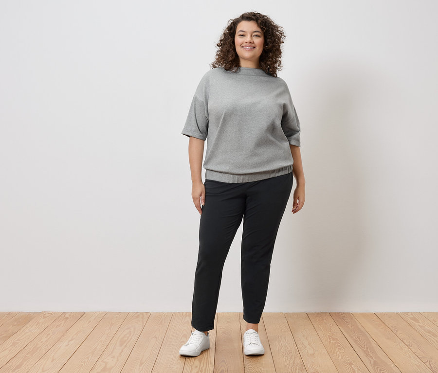 Une femme aux cheveux bouclés se tient devant un mur blanc, portant un sweat-shirt gris, un pantalon noir et des baskets blanches.