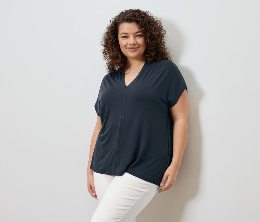 Une femme aux cheveux bouclés se tient devant un mur blanc, portant un t-shirt bleu marine et un pantalon blanc.