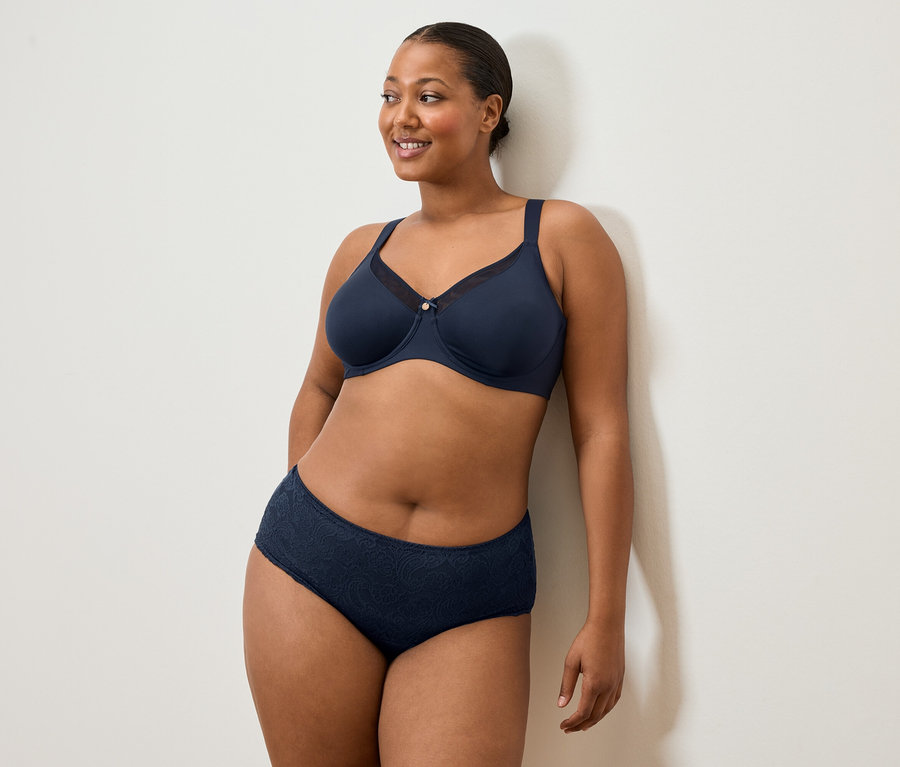 Femme en soutien-gorge et culotte bleu marine, adossée à un mur blanc, souriant.
