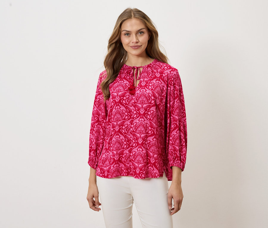 Femme portant un chemisier rose à motifs et un pantalon blanc.