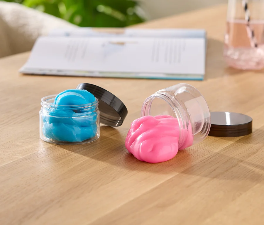 Deux pots de slime ouverts sur une table en bois ; l'un est droit avec du slime bleu, l'autre est couché sur le côté avec du slime rose qui en déborde. Un livre ouvert et un verre de liquide rose sont en arrière-plan.