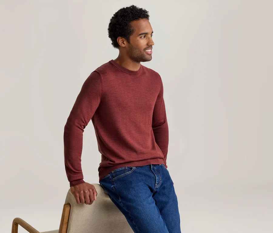 Homme en chemise rouge à manches longues et jean bleu, appuyé sur une chaise claire et souriant en regardant vers la droite.
