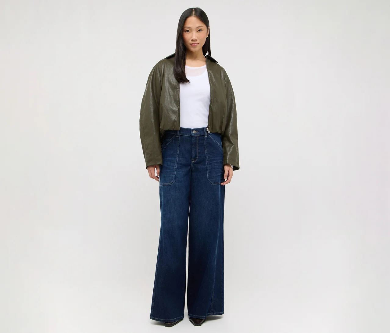 Une femme se tient de face, vêtue d'un t-shirt blanc, d'une veste courte vert olive et d'un jean large bleu foncé.