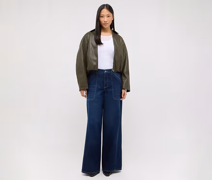 Une femme se tient de face, vêtue d'un t-shirt blanc, d'une veste courte vert olive et d'un jean large bleu foncé.