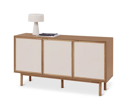 Un buffet en bois clair avec trois portes à panneaux crème, reposant sur quatre pieds. Une lampe blanche et une pile de magazines sont posées dessus.