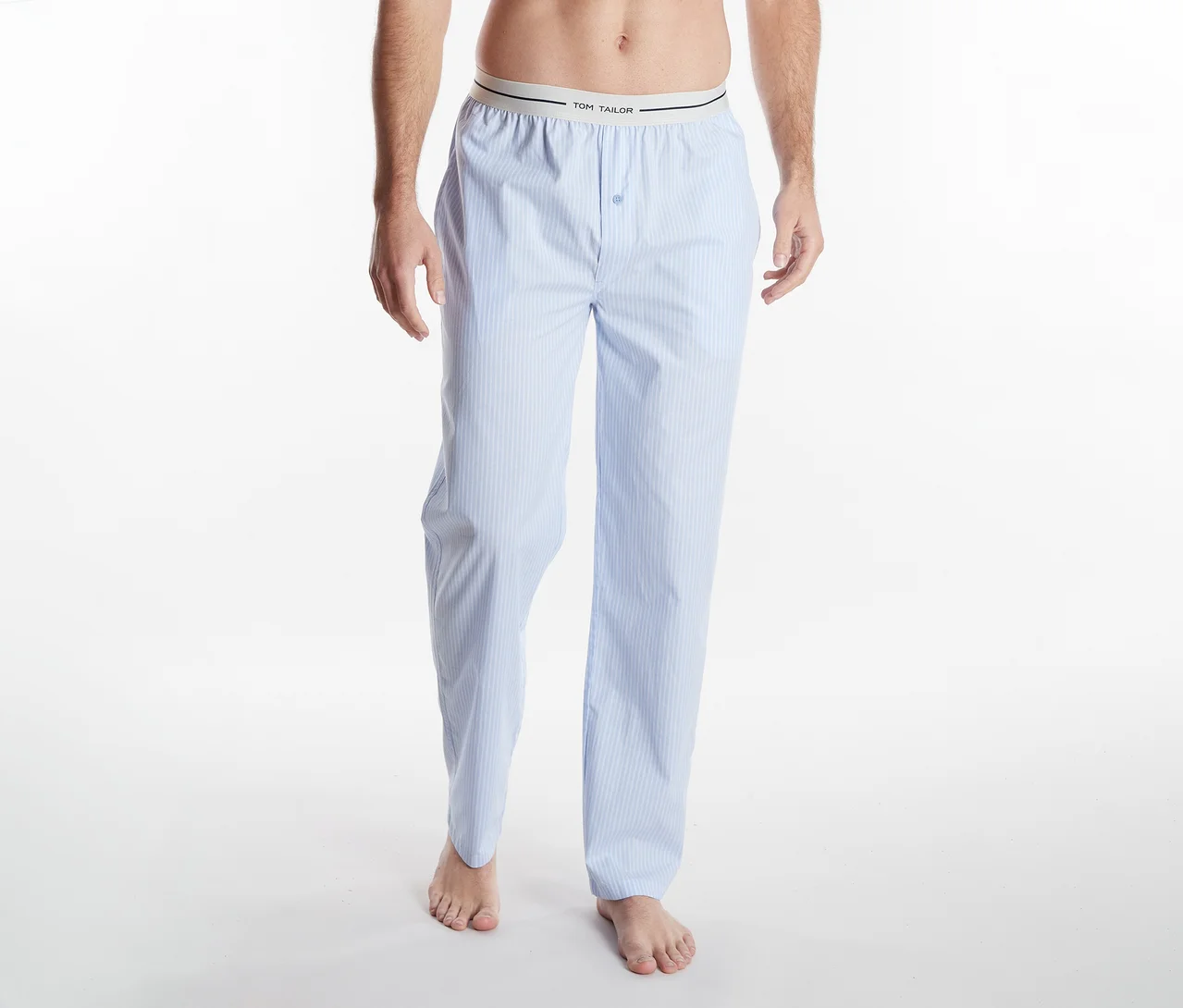 Une personne de la taille aux pieds, portant un pantalon de pyjama rayé bleu clair et blanc avec une ceinture "TOM TAILOR", pieds nus sur un fond blanc.