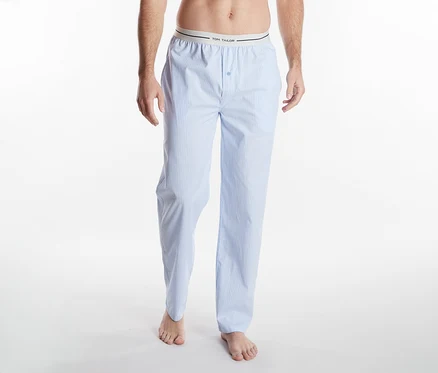 Une personne de la taille aux pieds, portant un pantalon de pyjama rayé bleu clair et blanc avec une ceinture "TOM TAILOR", pieds nus sur un fond blanc.